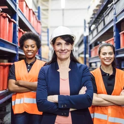 Drei selbstbewusst wirkende und lächelnde Frauen befinden sich in einem Lagerhaus. Eine trägt einen Schutzhelm.