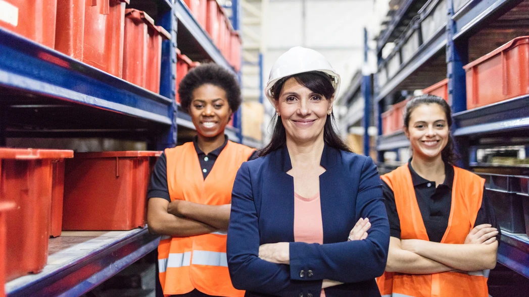Drei selbstbewusst wirkende und lächelnde Frauen befinden sich in einem Lagerhaus. Eine trägt einen Schutzhelm.