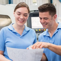 Zwei junge Fachkräfte in blauen Poloshirts betrachten einen technischen Plan vor einer Industriemaschine.