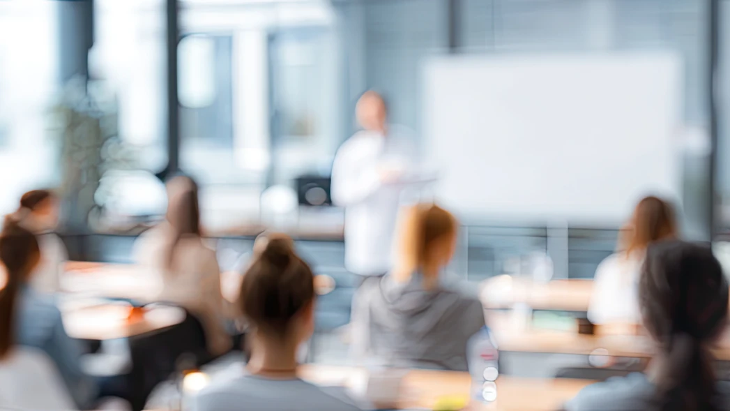 Verschwommene Aufnahme eines hellen Seminarraums mit Referenten und Teilnehmern vor einer Tafel