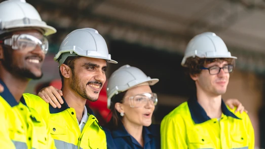 Vier lächelnde Fachkräfte in Warnwesten und mit Schutzhelmen. Symbolisiert Teamgeist und Sicherheit am Arbeitsplatz.